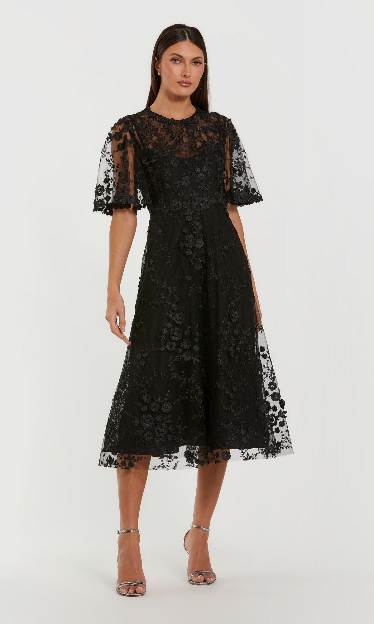 A woman poses in the Mac Duggal 69036 Midi Wedding Guest Dress in black, featuring floral-embroidered mesh and short flared sleeves, styled with silver strappy heels against a plain backdrop.